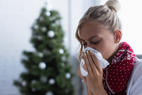 sick woman sneezing in paper napkin near christmas tree on blurred background