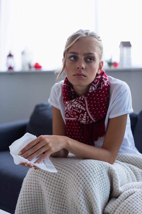diseased and pensive woman in warm scarf sitting on sofa with paper napkin
