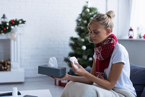 sick woman in warm scarf holding paper napkins near christmas tree on blurred background