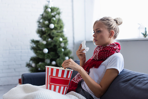 sick woman with paper napkin and bucket of popcorn watching tv on sofa near blurred christmas tree