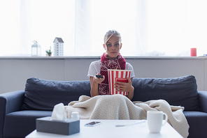 sick woman with remote controller and bucket of popcorn watching tv near blurred paper napkins and cup of warm drink