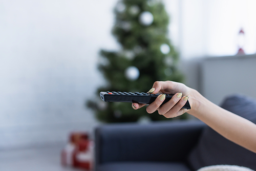 cropped view of female hand with remote controller near blurred christmas tree