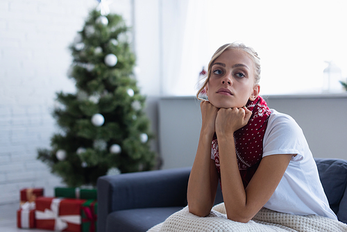 sick and sad woman looking away while sitting on sofa near blurred christmas tree