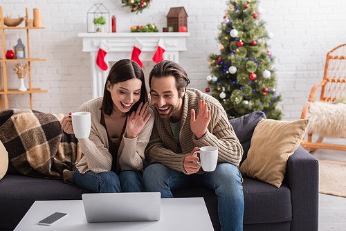 cheerful couple with tea cups waving hands during video call in decorated living room