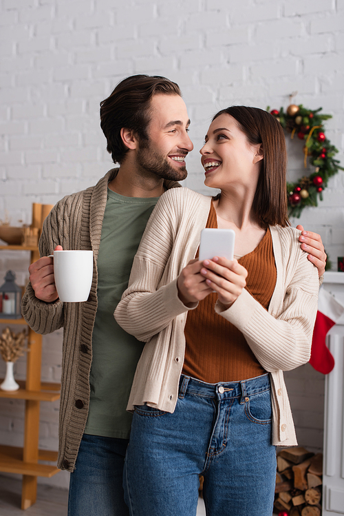 smiling woman using smartphone near husband with cup of tea