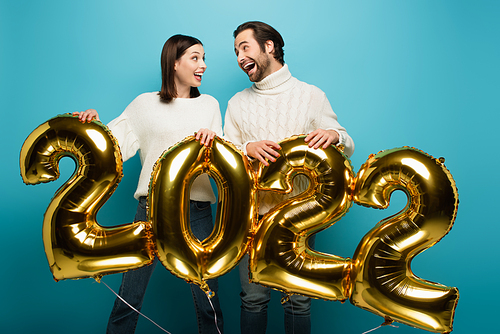 amazed couple in warm sweaters looking at each other near golden balloons in shape of 2022 on blue