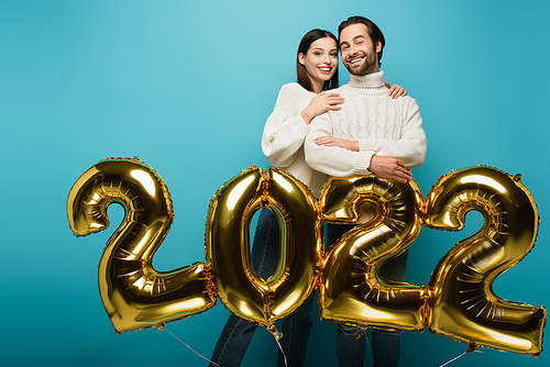 happy woman embracing smiling man near golden balloons in shape of 2022 on blue