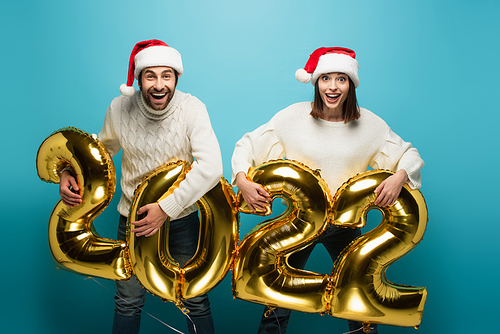 excited couple in santa hats holding golden balloons in shape of 2022 on blue