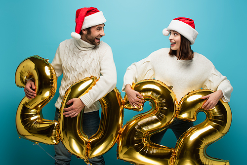 astonished couple in santa hats looking at each other near golden balloons in shape of 2022 on blue