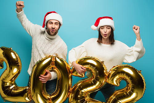 excited couple in santa hats holding golden balloons in shape of 2022 and showing win gesture on blue