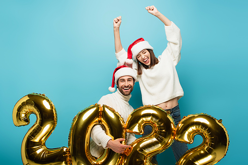 happy man in santa hat  near golden 2022 balloons and woman showing win gesture on blue