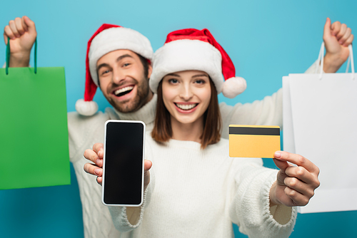 woman in santa hat showing credit card and smartphone near blurred man with shopping bags isolated on blue