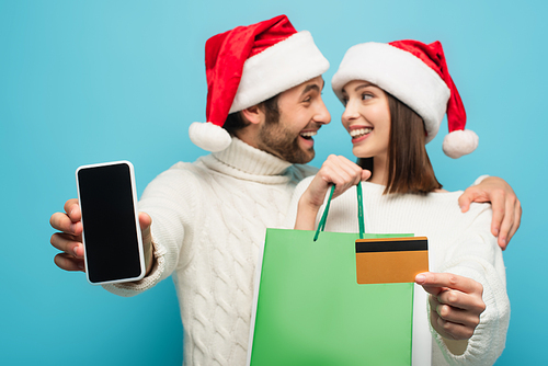 blurred couple in santa hats smiling at each other while holding smartphone, credit card and shopping bags isolated on blue