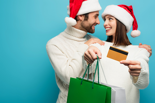 blurred couple in santa hats smiling at each other near shopping bags and credit card isolated on blue