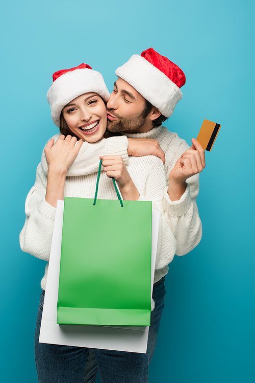 man in santa hat kissing happy woman holding credit card and shopping bags isolated on blue