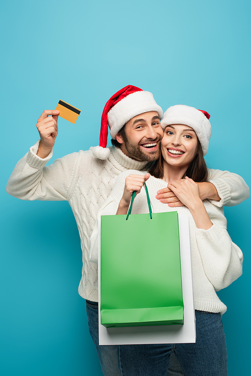 happy man in santa hat showing credit card and embracing woman with shopping bags on blue