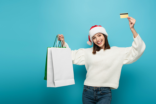 cheerful woman in santa hat showing shopping bags and credit card on blue