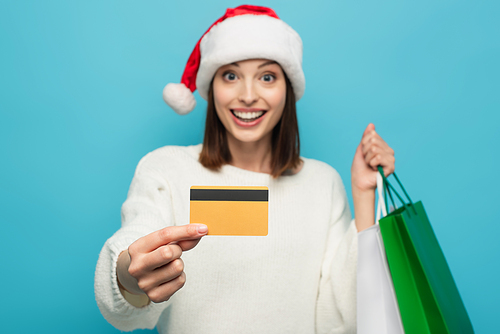 blurred woman smiling while holding credit card and shopping bags isolated on blue