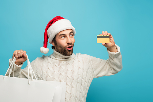 amazed man in santa hat looking at credit card while holding purchases isolated on blue