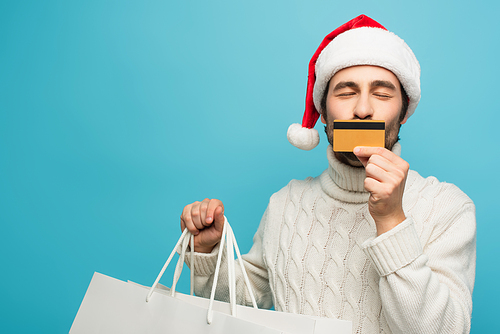 man in santa hat kissing credit card while holding shopping bags isolated on blue