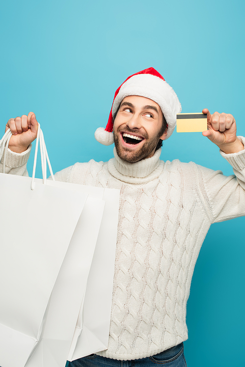 astonished man in santa hat holding purchases and credit card isolated on blue