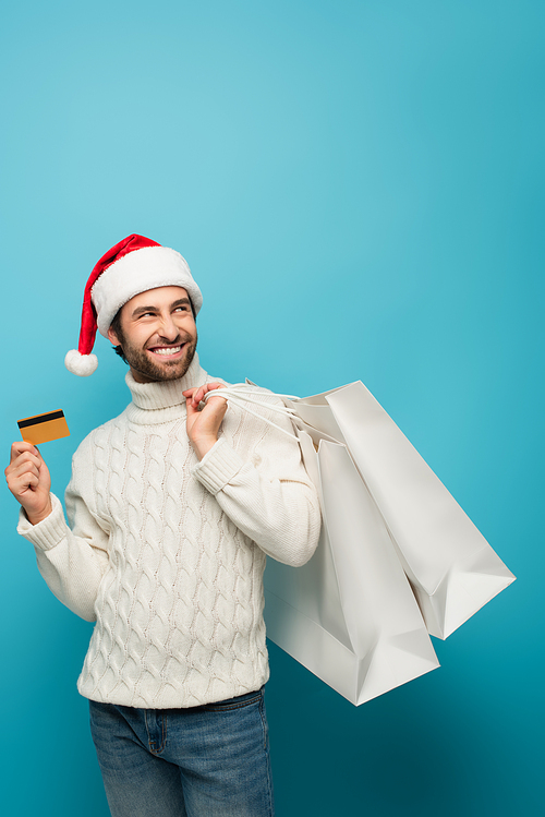 pleased man in santa hat holding credit card and shopping bags while looking away on blue