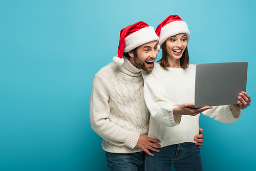 amazed couple in warm sweaters and santa hats looking at laptop on blue