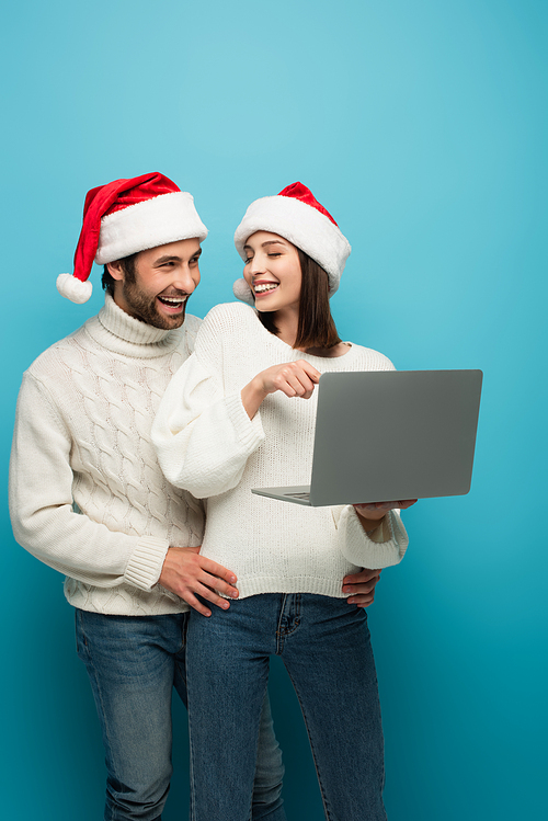 cheerful woman pointing at laptop near smiling man in santa hat on blue