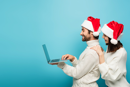 side view of happy couple in santa hats and warm sweaters looking at laptop on blue