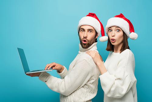 astonished couple in santa hats  near laptop on blue