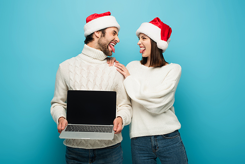 cheerful couple in santa hats sticking out tongues near laptop with blank screen on blue