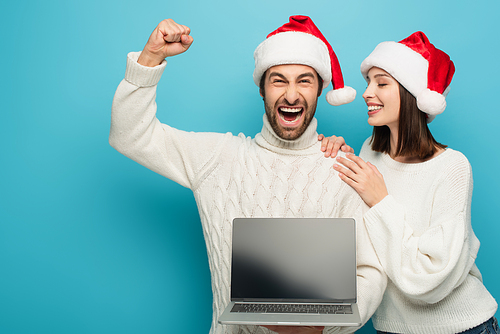 excited man showing win gesture while holding laptop near happy woman in santa hat on blue