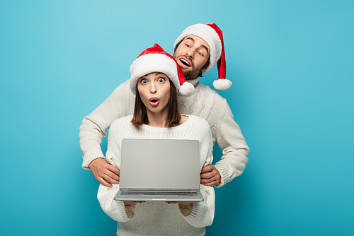 curious man in santa hat looking at laptop near amazed woman on blue