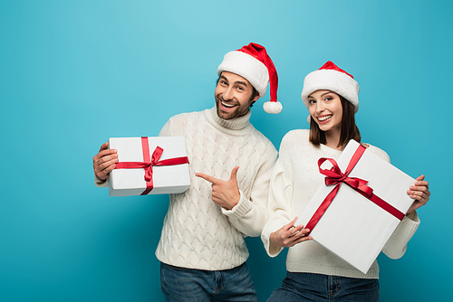 cheerful man in santa hat pointing at gift box near happy woman with present on blue