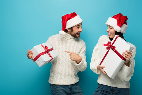 amazed man in santa hat pointing at gift box near woman sticking out tongue on blue