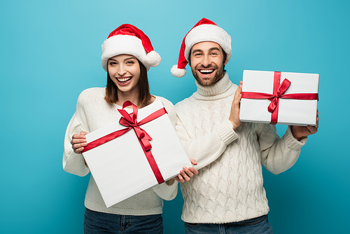 joyful couple in white sweaters and santa hats holding presents and  on blue