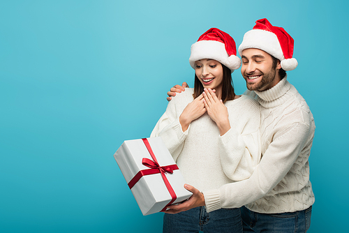 smiling man in santa hat holding christmas gift near amazed woman isolated on blue