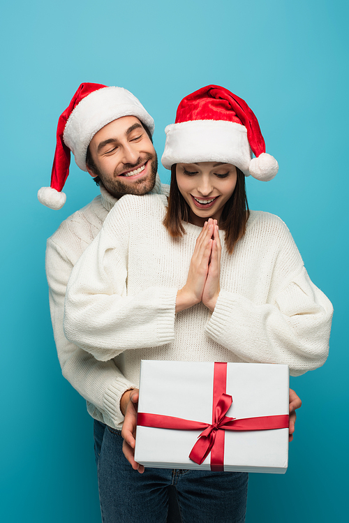 smiling man in santa hat holding present near amazed woman isolated on blue