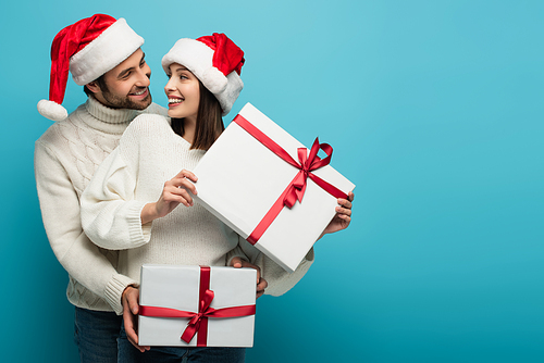 pleased couple in santa hats holding christmas gifts while looking at each other on blue