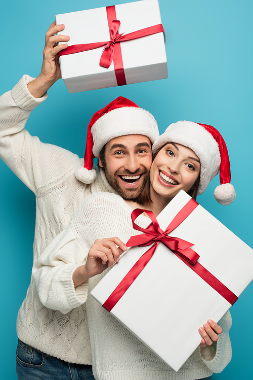 cheerful couple in santa hats and white sweaters holding christmas presents on blue