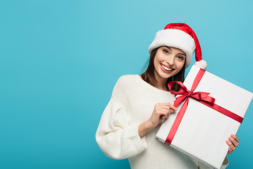 joyful woman in santa hat  while holding present isolated on blue