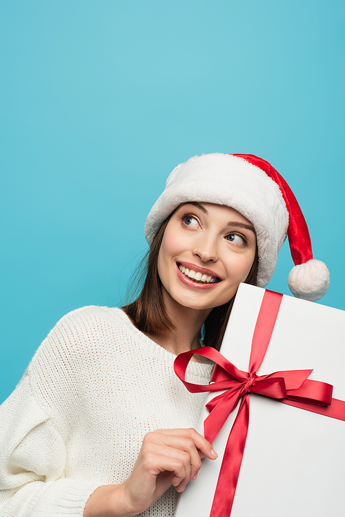 cheerful woman in santa hat looking away while holding christmas present isolated on blue