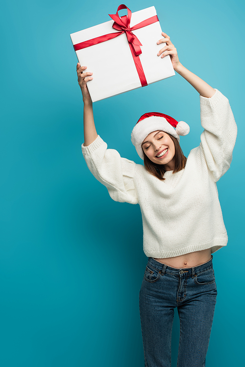 happy woman in white sweater and santa hat holding gift box in raised hands on blue