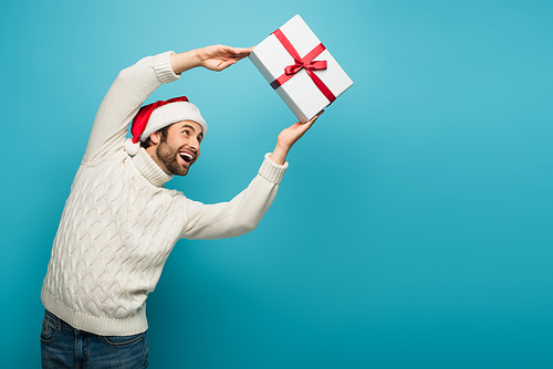 excited man in santa hat and warm sweater catching gift box on blue