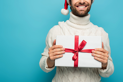 cropped view of smiling man in santa hat and white sweater holding present isolated on blue