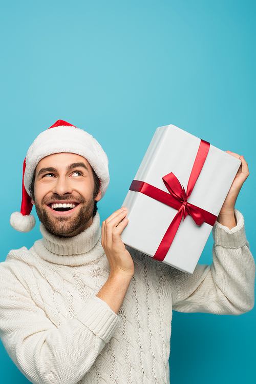 happy and curious man in santa hat shaking gift box isolated on blue