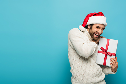 cheerful man in white sweater and santa hat showing present on blue