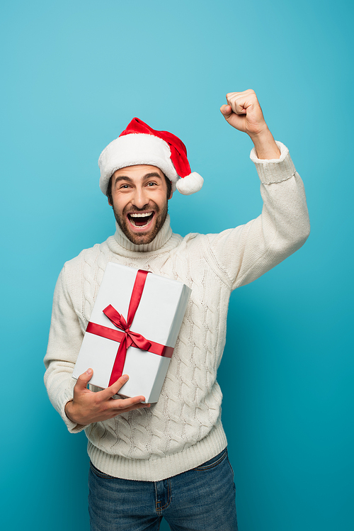 excited man in santa hat holding gift box and showing yeah gesture on blue