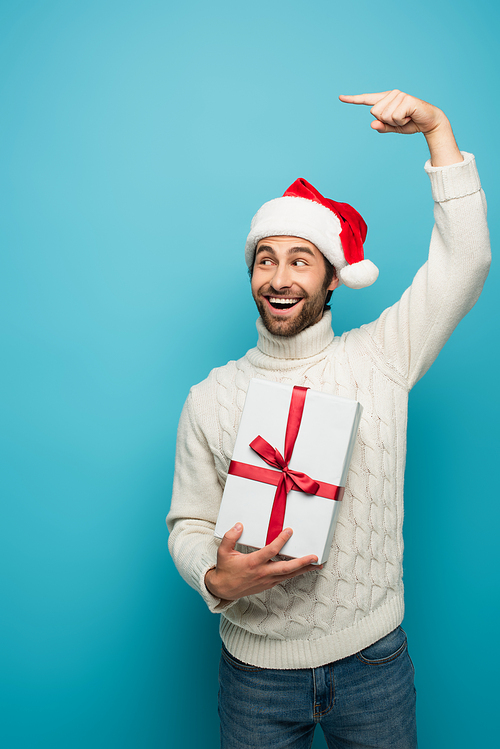 cheerful man in santa hat looking away and pointing with finger while holding gift isolated on blue