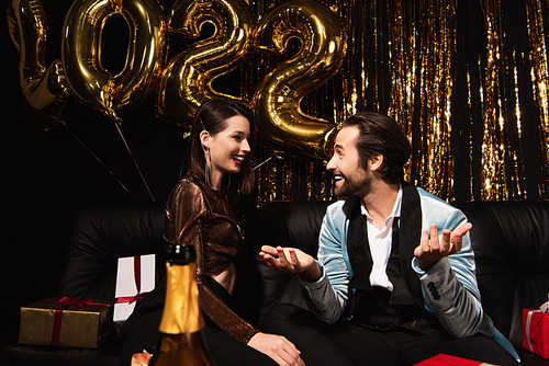 smiling man talking to elegant woman near christmas decoration and balloons in shape of 2022 on black